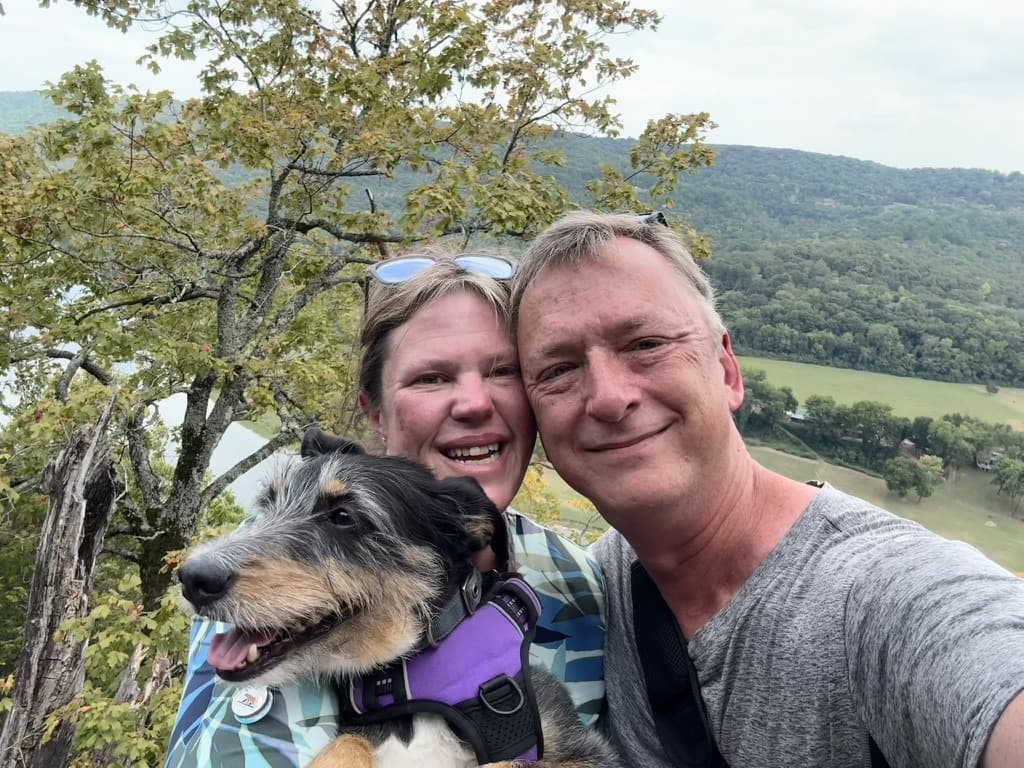 Jason and Trisha outdoors with their dog on a scenic hilltop